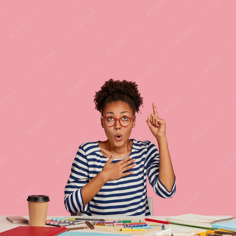 Frightened emotional dark skinned female designer raises index finger, keeps hand on breast, points upwards, notices unbelievable item, uses notebook, caryons for making sketch sits in co working space Free Photo