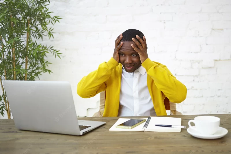 Frustrated dark skinned businessman sitting at workplace with hands on head, feeling stressed, looking at laptop screen in panic, unable to keep company afloat, having not enough money to run business Free Photo