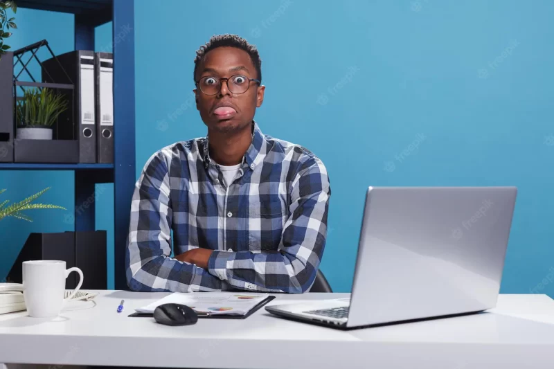 Funny and goofy office working person making amusing playful face while sticking out tongue. childish and dumb young adult man with dullard face expression while in company workspace. Free Photo