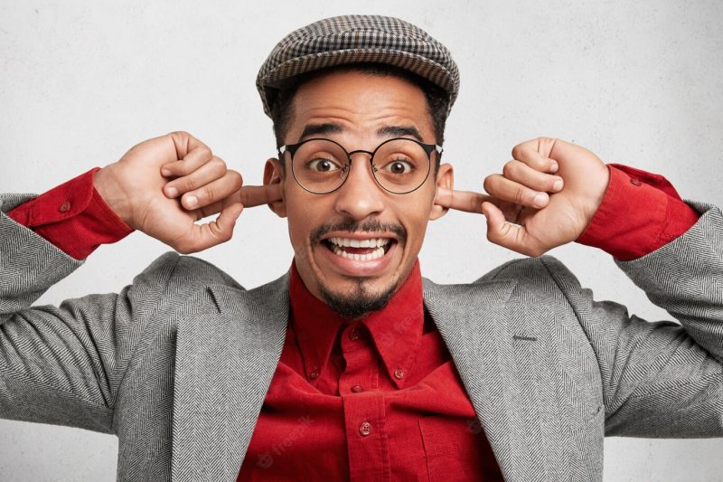 Funny mixed race man wears cap and red shirt with jacket, plugs ears and smiles joyfully Free Photo