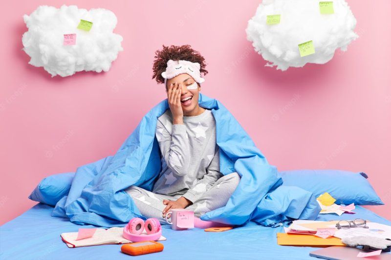 Girl laughs happily makes face palm wears soft pajama and blindfold works distantly on quarantine poses with papers sticky notes on bed stays at home alone Free Photo