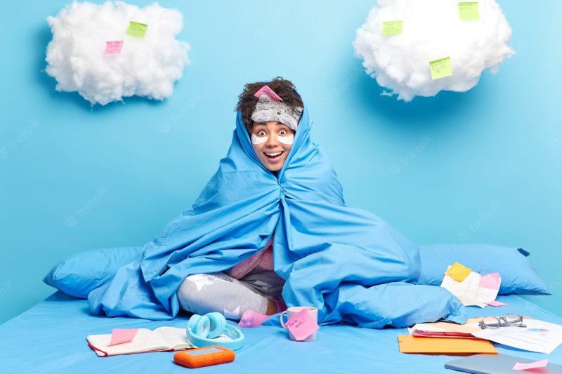 Girl studies remotely from home during quarantine wrapped in blanet makes mlist to do on sticky notes looks happily isolated on blue wall Free Photo