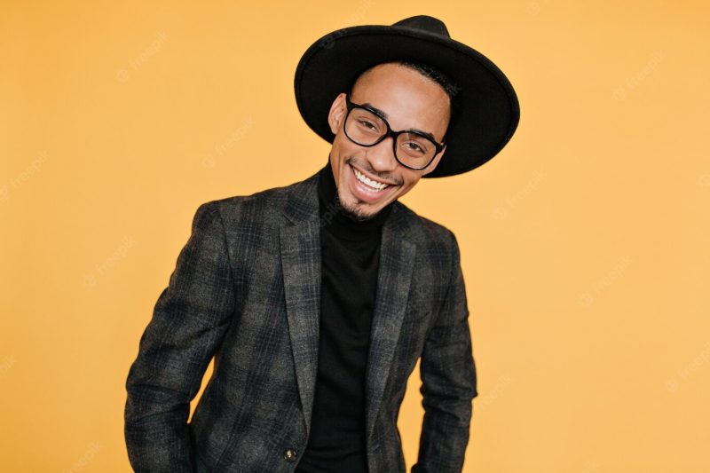 Glad african man in black shirt and gray jacket posing. photo of positive mulatto guy with sincere smile isolated on orange wall. Free Photo