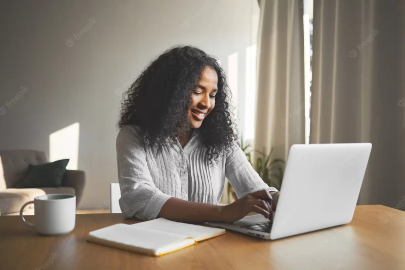 Gorgeous positive young dark skinned female blogger keyboarding on generic laptop, smiling, being inspired while creating new content for her travel blog, sitting at desk with diary and mug Free Photo