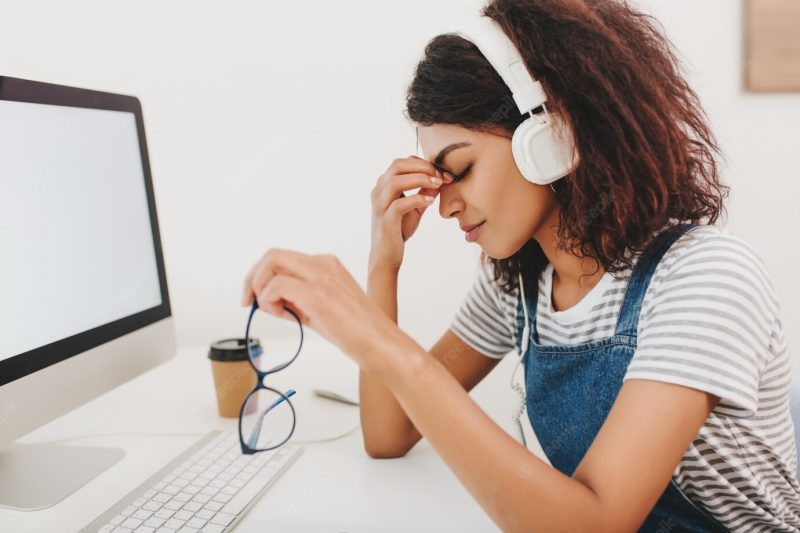 Graceful black woman in headphones with headache sitting beside computer and holding glasses in hand Free Photo