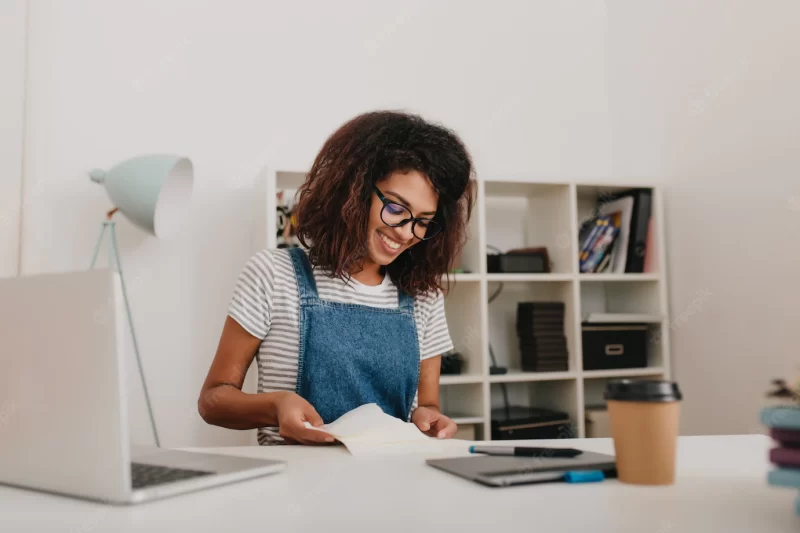 Graceful girl wih curly brown hair reading documents with pretty smile sitting in her office Free Photo