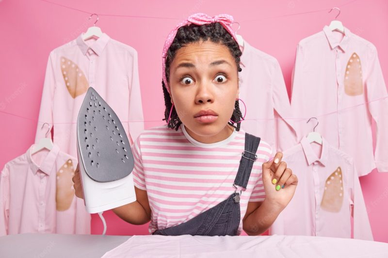 Greatly surprised ethnic woman with braids stares shocked at camera busy ironing at home holds electric irond dressed in casual clothes does daily domestic chores stands against ironed shirts Free Photo