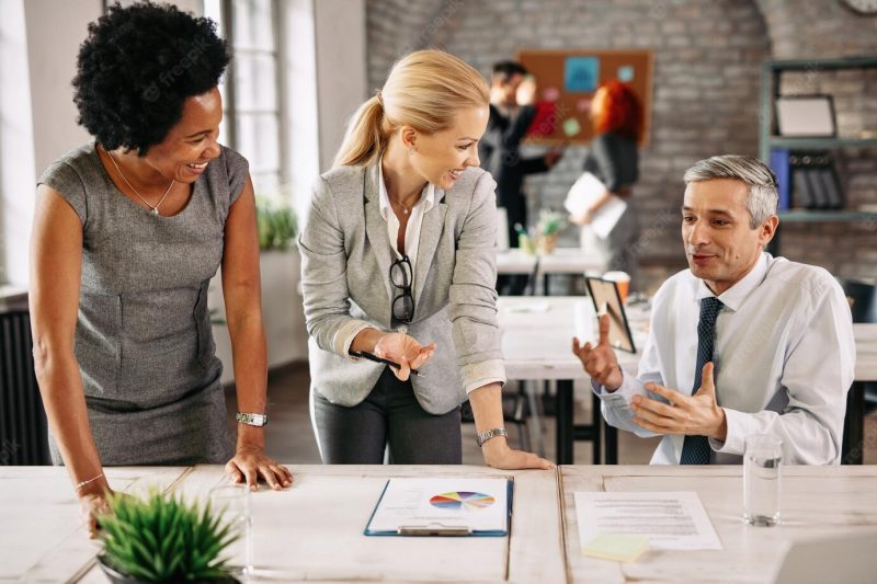 Group of smiling business people analyzing reports and communicating while working together in the office there are people in the background Free Photo