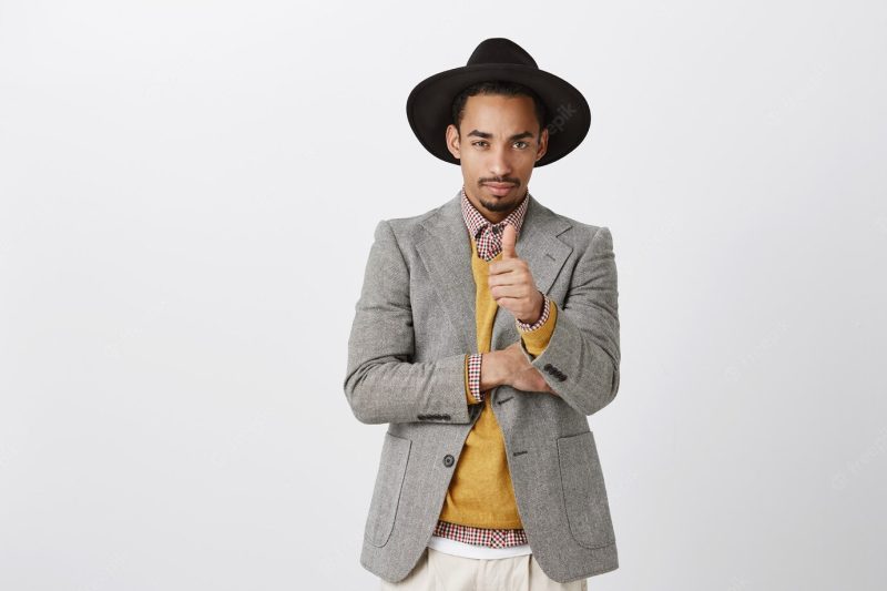 Guy came to make business. portrait of serious good-looking African entrepreneur in stylish black hat and jacket, showing thumb up, being focused while discussing new concept of work in office Free Photo