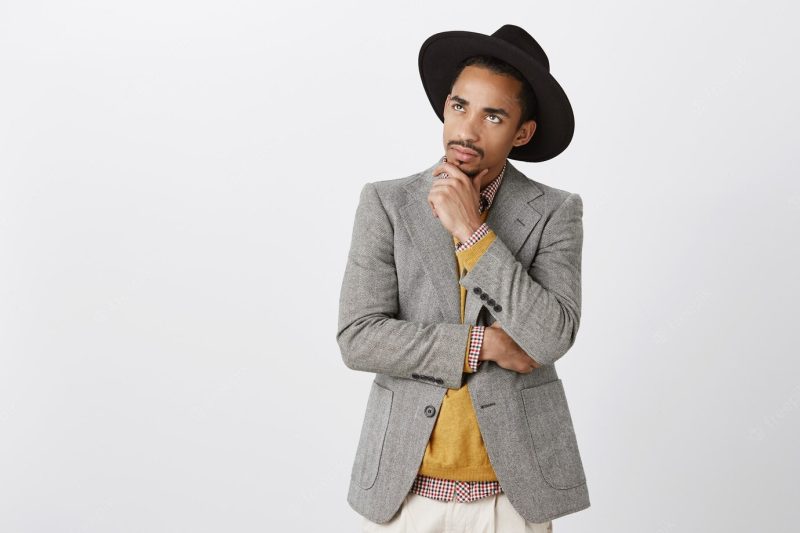 Guy focused on thoughts, making hard decision. concentrated serious dark-skinned male freelancer in black hat and stylish jacket, touching chin, looking up with concerned thoughtful expression Free Photo