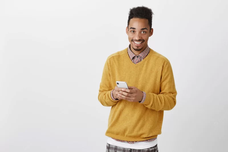 Guy wants to make call. indoor shot of pleased handsome African-American male model with Afro haircut in yellow sweater, holding smartphone, smiling broadly while messaging with friend Free Photo