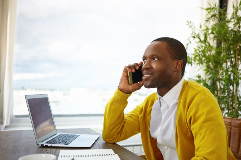 Half profile shot of handsome stylish Afro American designer talking on mobile phone to client, discussing details and ideas of house interior project, having inspired look, sitting by window Free Photo