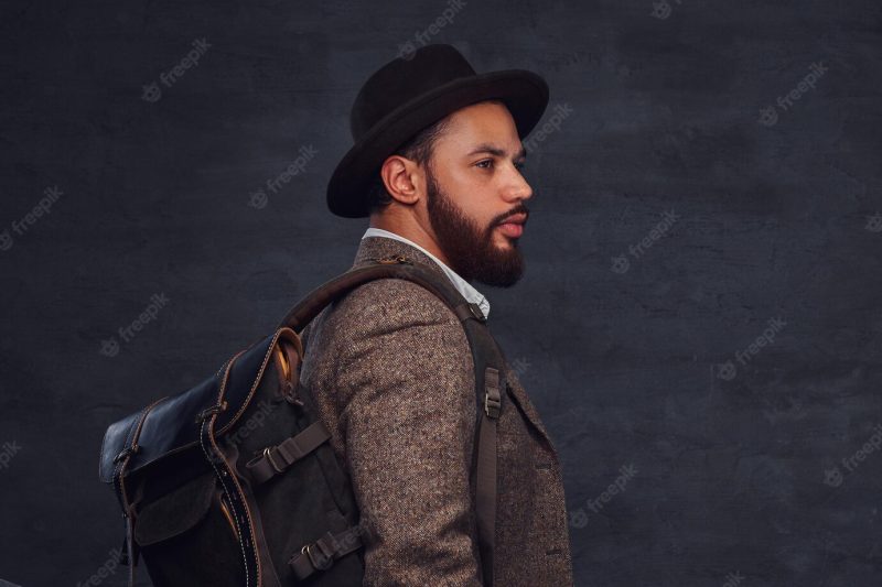 Handsome afro-american traveler in a brown jacket and hat with the backpack, stands in a studio. isolated on a dark background. Free Photo