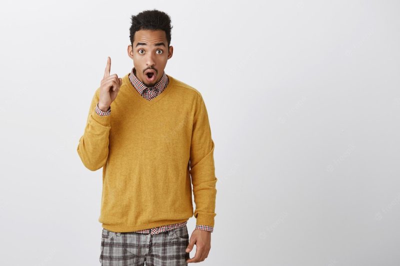 Handsome guy dropping jaw from incredible news. portrait of emotive young african-american male with afro hairstyle in trendy yellow sweater pointing up, looking with shock and wonder over gray wall Free Photo