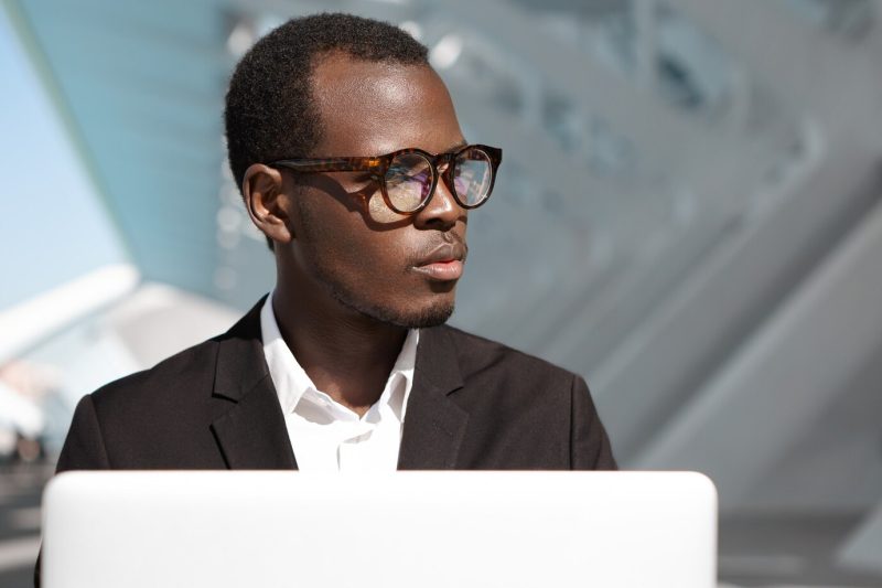 Handsome successful young Afro American corporate worker in eyewear and black suit sitting outdoors in front of laptop pc, looking away, having thoughtful expression, absorbed in business issues Free Photo