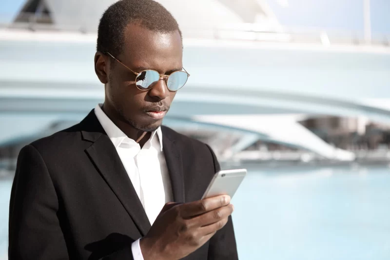 Handsome young African American office worker in elegant black suit and eyewear standing in urban surroundings, looking concentrated while trying to call cab using online app on his mobile phone Free Photo