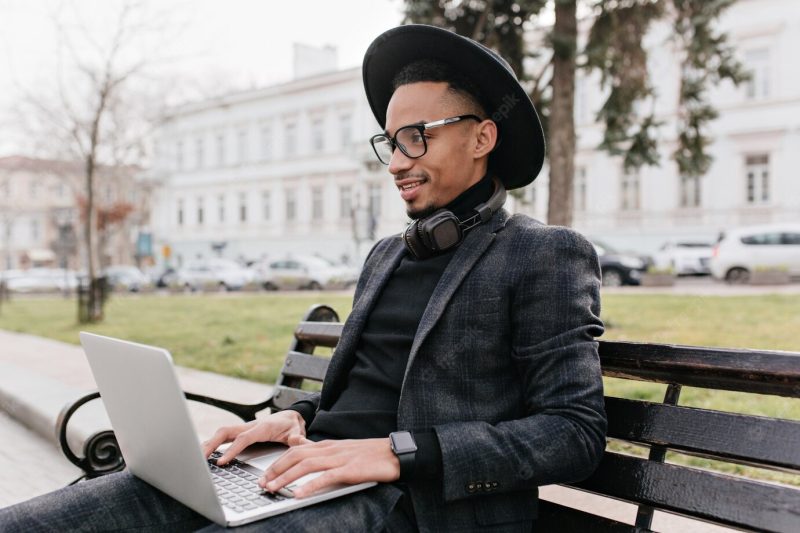 Handsome young freelancer working with computer in park. outdoor portrait of glad african guy in hat studying with laptop on bench. Free Photo