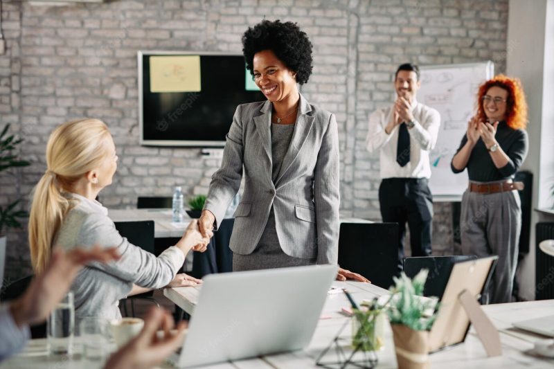 Happy african american businesswoman shaking hands with her female colleague in the office while other coworkers are applauding them Free Photo