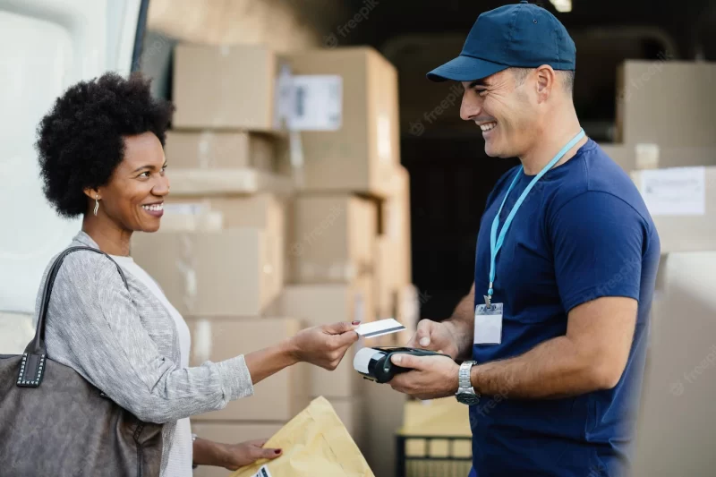 Happy African American woman using credit card while making contactless payment to a courier for package delivery Free Photo