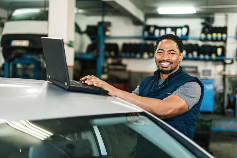 Happy black auto mechanic working on a computer and looking at camera in repair shop Free Photo