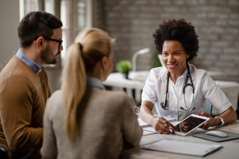 Happy black female doctor talking to a couple and showing them medical test results on a touchpad during consultations at clinic Free Photo