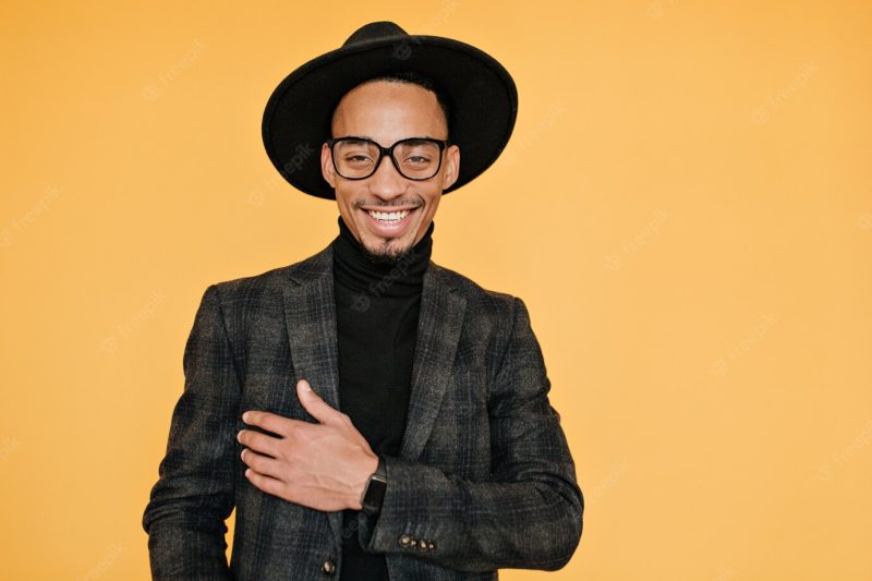 Happy black young man wears elegant dark suit posing with pleased smile. indoor photo of relaxed mulatto male model in glasses enjoying photo shoot. Free Photo