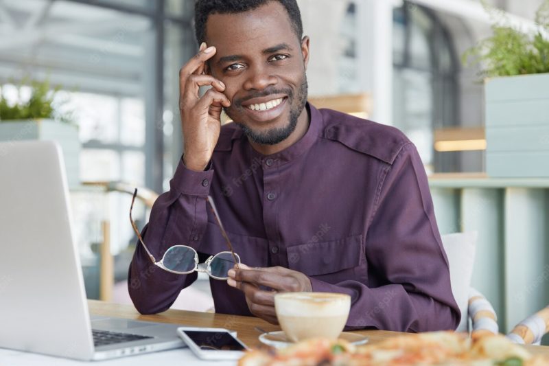 Happy dark skinned male freelancer uses modern electronic gadgets for remote work, sits against cozy coffee shop interior, drinks aromatic espresso Free Photo