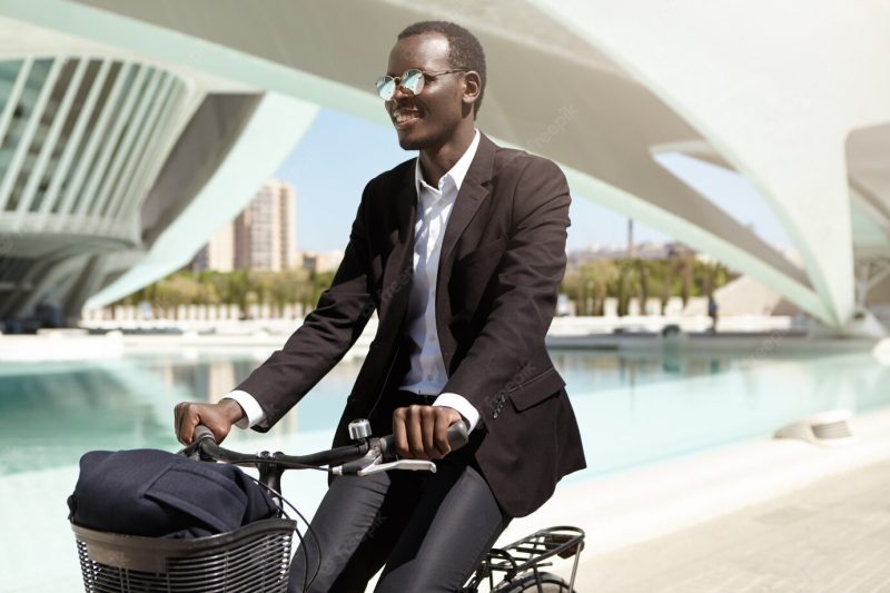 Happy environmentally conscious African American employee wearing black formal suit and sunglasses choosing bicycle over public transport or car to get to office, enjoying ride in urban setting Free Photo