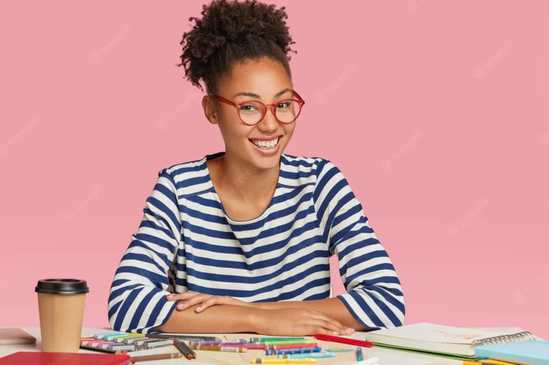 Happy ethnic student designer draws sketch for university exam, wears striped jacket and glasses Free Photo