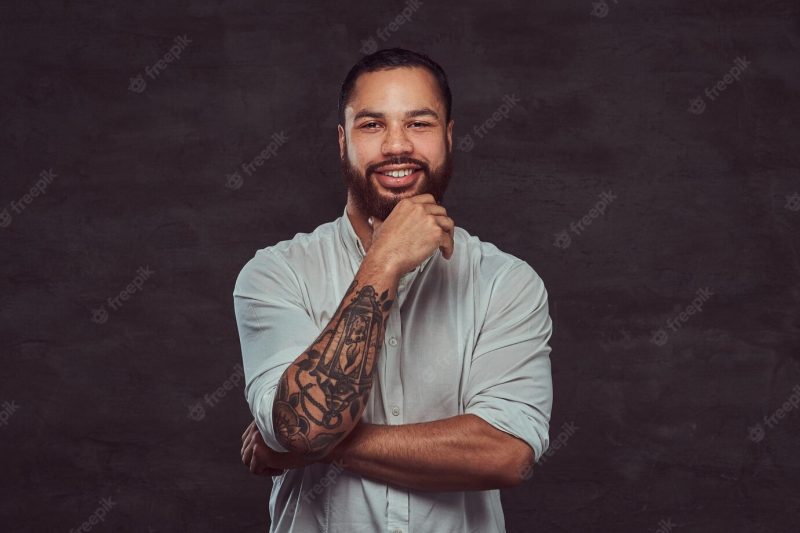 Happy handsome african-american tattooed man with stylish hair and beard in a white shirt, holding hand on his chin. Free Photo