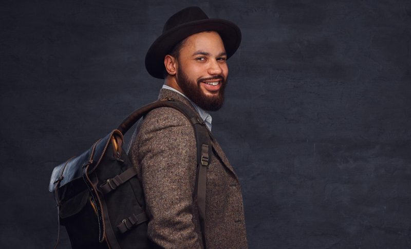 Happy handsome afro-american traveler in a brown jacket and hat with the backpack, stands in a studio. isolated on a dark background. Free Photo