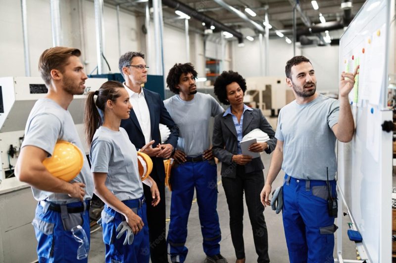 Happy manual worker presenting the results of business development while giving presentation to company leaders and his team in a factory Free Photo