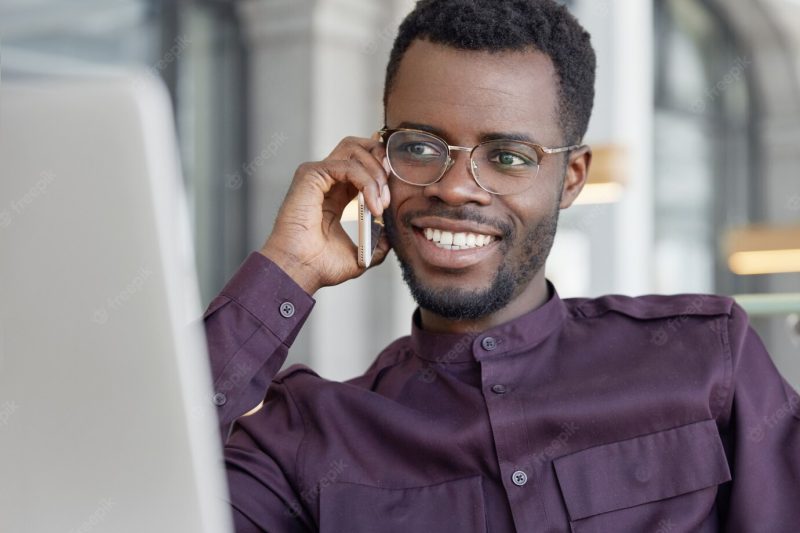 Happy middle aged african male business worker has pleasant talk with friend via smart phone, shares success in high sales Free Photo