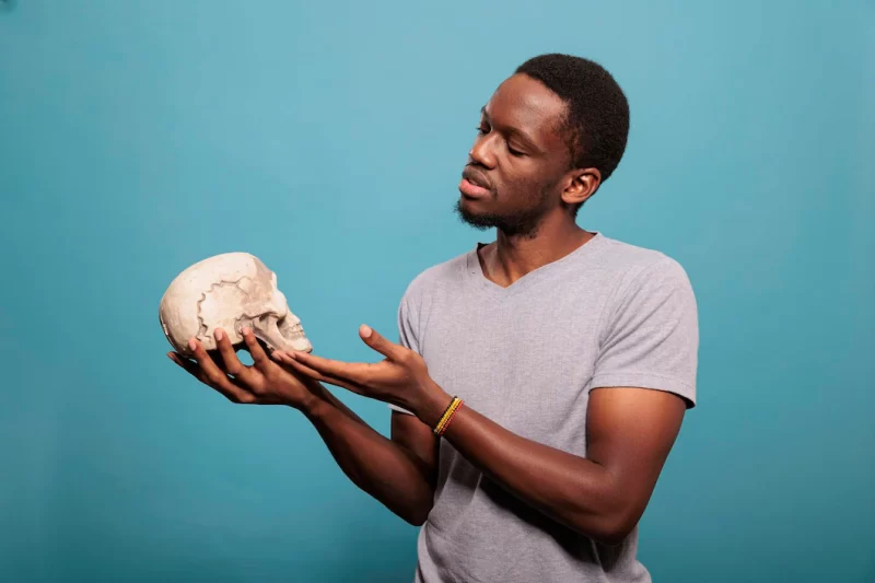 Happy model holding skull from human skeleton in hand, standing over blue background to learn genetic knowledge. scientific guy studying biology and anatomy subject made of bones. Free Photo