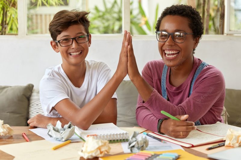 Happy young partners cooperate together in workspace, give high five to each other, have overjoyed expressions, work on research, write down records in notepad, ponder on idea, work as team. Free Photo