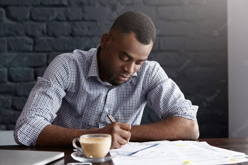 Hardworking young African office worker handwriting, filling in papers while doing accounting Free Photo