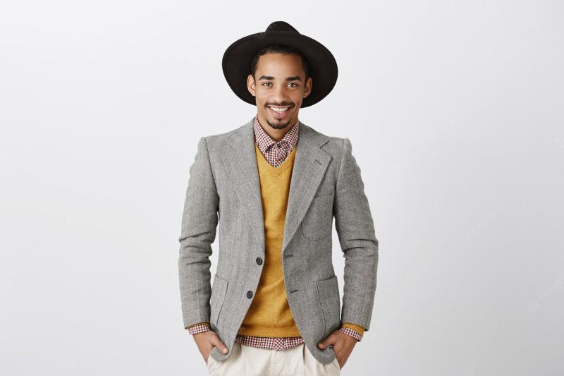 He knows how to make great deals. portrait of self-assured handsome male model with dark skin, wearing trendy jacket and hat, holding hands in pockets and smiling confidently Free Photo