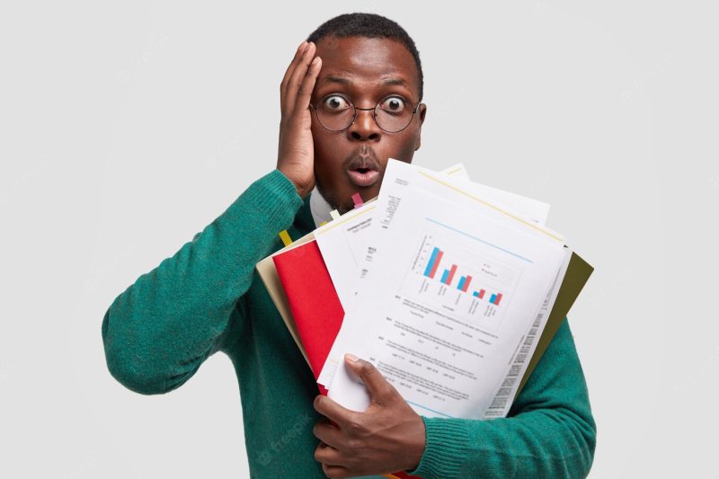 Headshot of puzzled African American man touches head, stares with bugged eyes, opens mouth from amazement, holds paper documents Free Photo