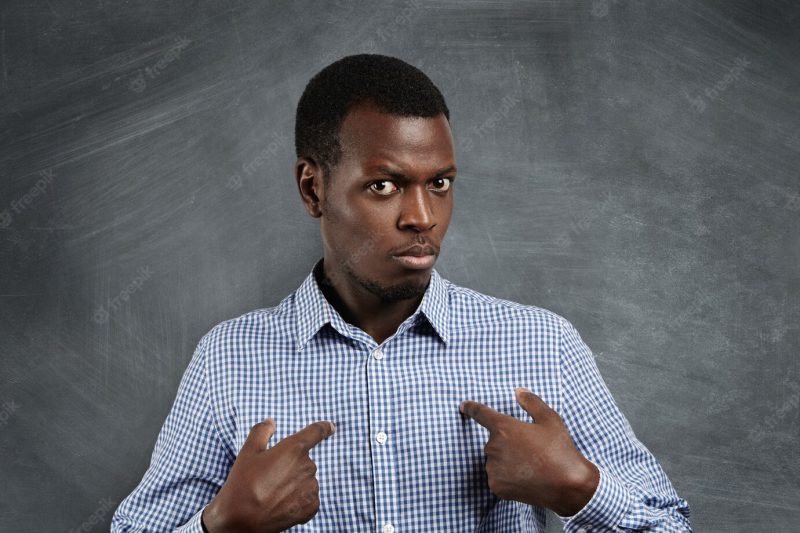 Headshot of young dark-skinned man pointing at himself, making excuses or verbally defending, having perplexed and puzzled look as if saying: who me? human face expressions, emotions and feelings Free Photo