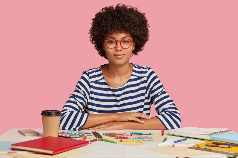 Photo of concentrated relaxed dark skinned young woman makes okay gesture with both hands, meditates at workplace, feels calm and relaxed, dressed in striped clothes, isolated on pink, draws picture Free Photo