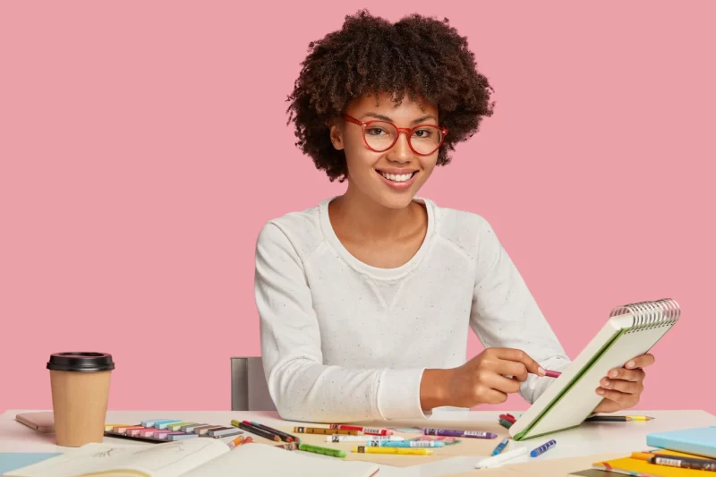 Horizontal shot of cheerful Afro African painter wears red rim spectacles, makes sketches in spiral notebook with pencil, enjoys aromatic beverage, surrounded with stationary, smiles positively Free Photo