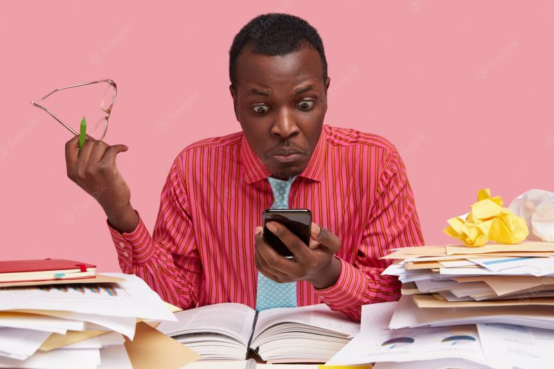 Horizontal shot of emotive stupefied puzzled dark skinned male worker holds mobile phone, stares at screen, receives message for paying bills Free Photo