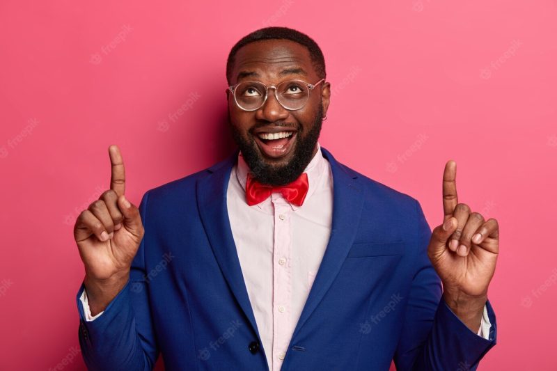 Horizontal shot of happy unshaven afro american office worker points at copy space above, shows something on blank wall, dressed elegantly Free Photo