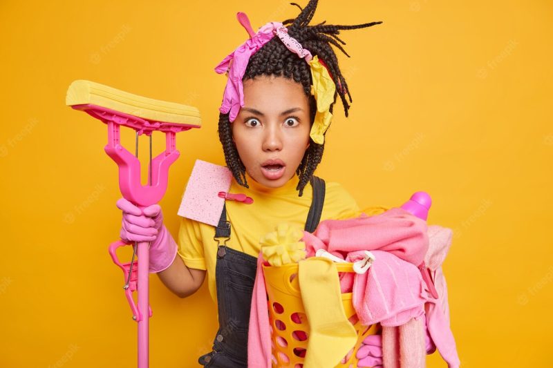 Horizontal shot of stunned housewife with dreadlocks has rubber gloves on head stares shocked Free Photo