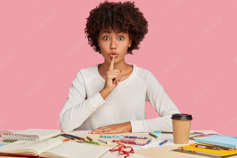 Horizontal shot of surprised dark skinned female illustrator keeps index finger over mouth, demonstrates silence gesture, sits at desktop with notepad, colored pencils, isolated over pink wall Free Photo