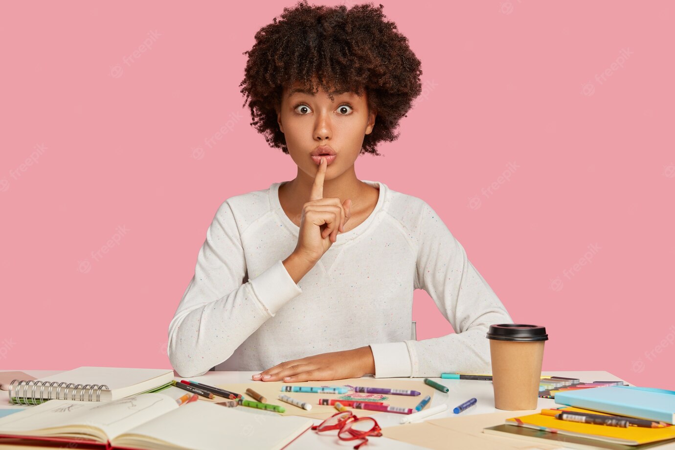 Horizontal Shot Surprised Dark Skinned Female Illustrator Keeps Index Finger Mouth Demonstrates Silence Gesture Sits Desktop With Notepad Coloured Pencils Isolated Pink Wall 273609 29531