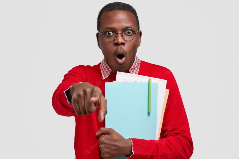 Horizontal shot of surprised scared black man points at camera, opens mouth from shock, carries textbooks closely, wears round spectacles Free Photo