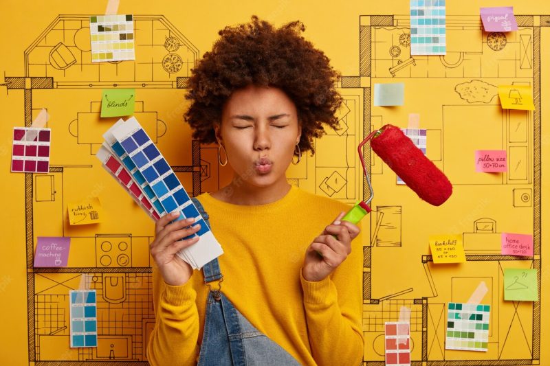 Housewife keeps lips rounded, busy with house renovation, holds paint brush and color palette, makes repair in apartmet according to design project. house painter poses over sketch on yellow wall Free Photo