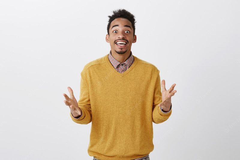 How could you miss this awesome party. portrait of overwhelmed attractive African guy with Afro haircut in yellow sweater gesturing with spread palms, talking with excitement about favorite singer Free Photo