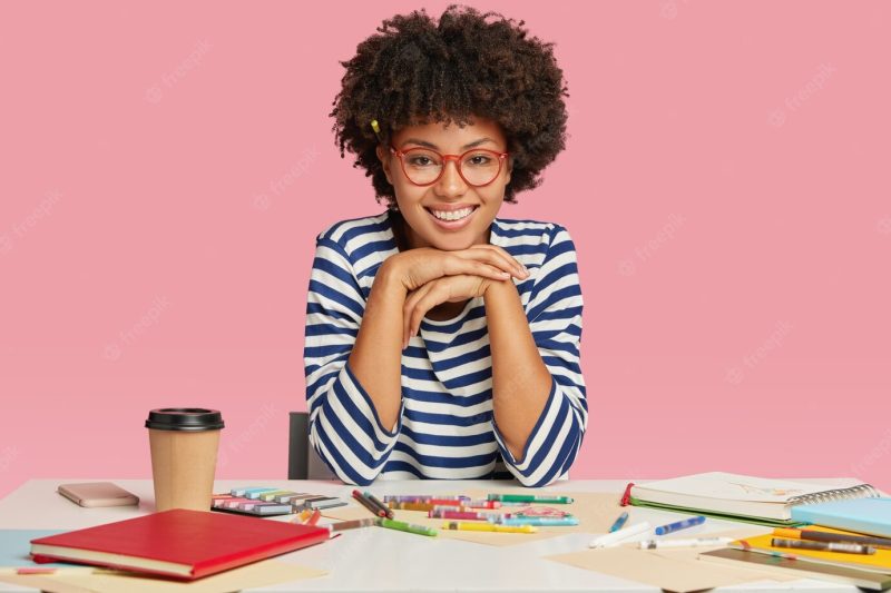 Image of happy teenage girl with toothy smile, holds hands under chin, rejoices positive comments about her work Free Photo